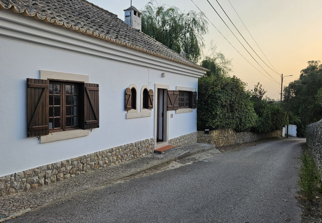 Casa rústica na beira de uma estrada rural, com paredes brancas e janelas tradicionais. 