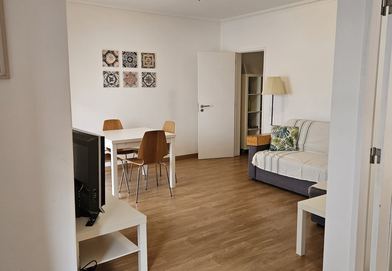 Sala de estar aconchegante com sofá, mesa de jantar para quatro pessoas e TV em móvel branco, iluminada por luz natural.