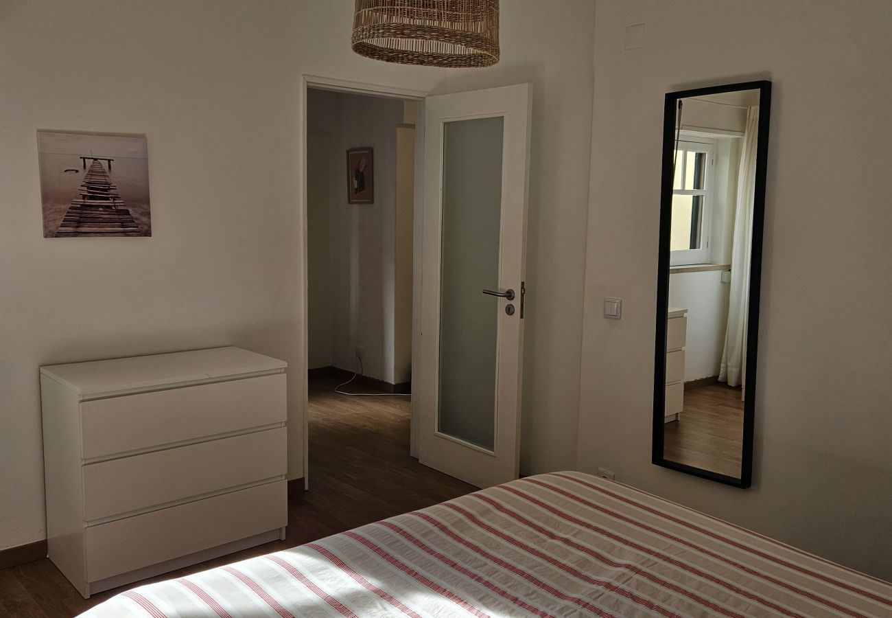 Quarto iluminado com cama de casal, cômoda branca, espelho de parede e luminária de vime, proporcionando um ambiente acolhedor e elegante.