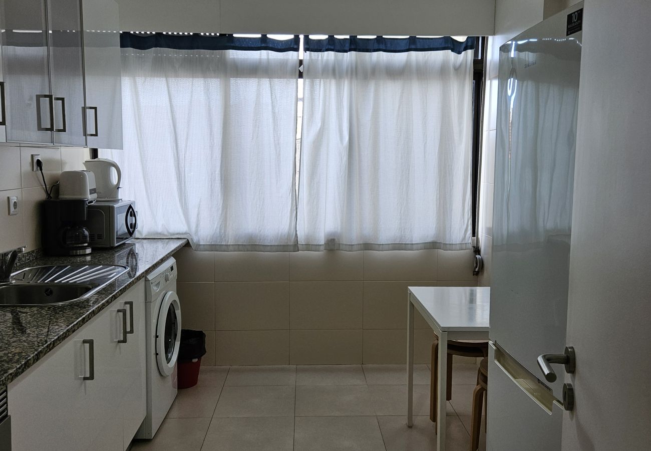 Cozinha funcional com armários brancos, bancada de granito, eletrodomésticos modernos e mesa para refeições, iluminada por uma grande janela.