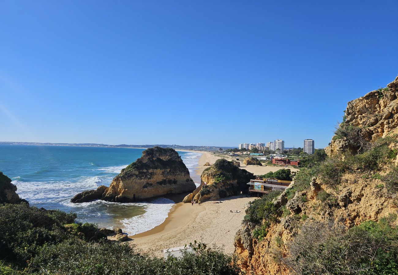 Vista panorâmica de uma praia dourada cercada por falésias imponentes e vegetação verde, com o mar azul ao fundo e edifícios à distância.