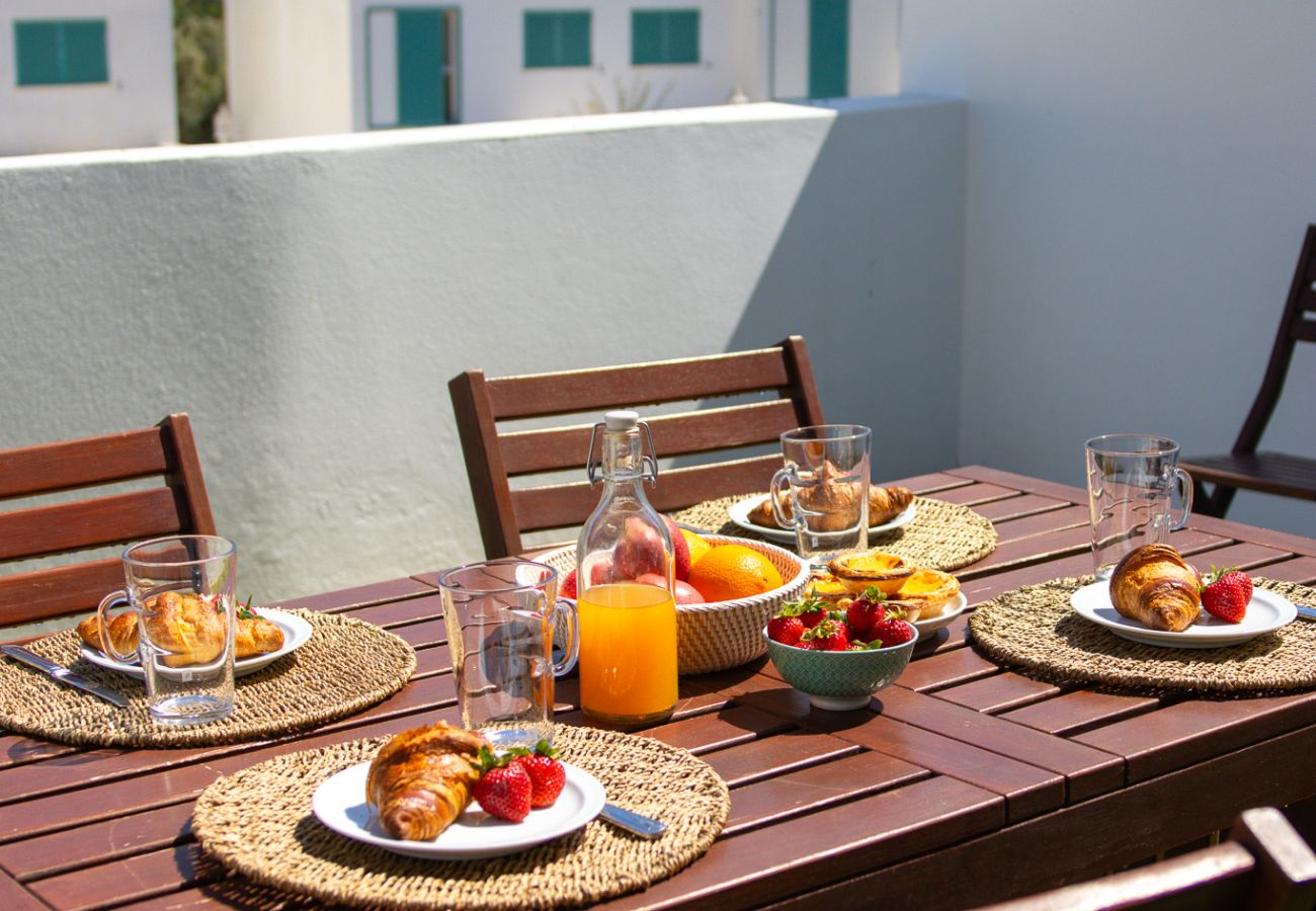 Mesa de jantar de madeira ao ar livre, posta para o café da manhã com croissants, suco de laranja, frutas e taças de vidro, com um fundo de casas bran