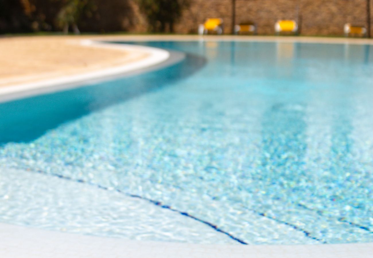 Piscina cristalina ao ar livre com espreguiçadeira, óculos escuros e roupa de banho no primeiro plano.