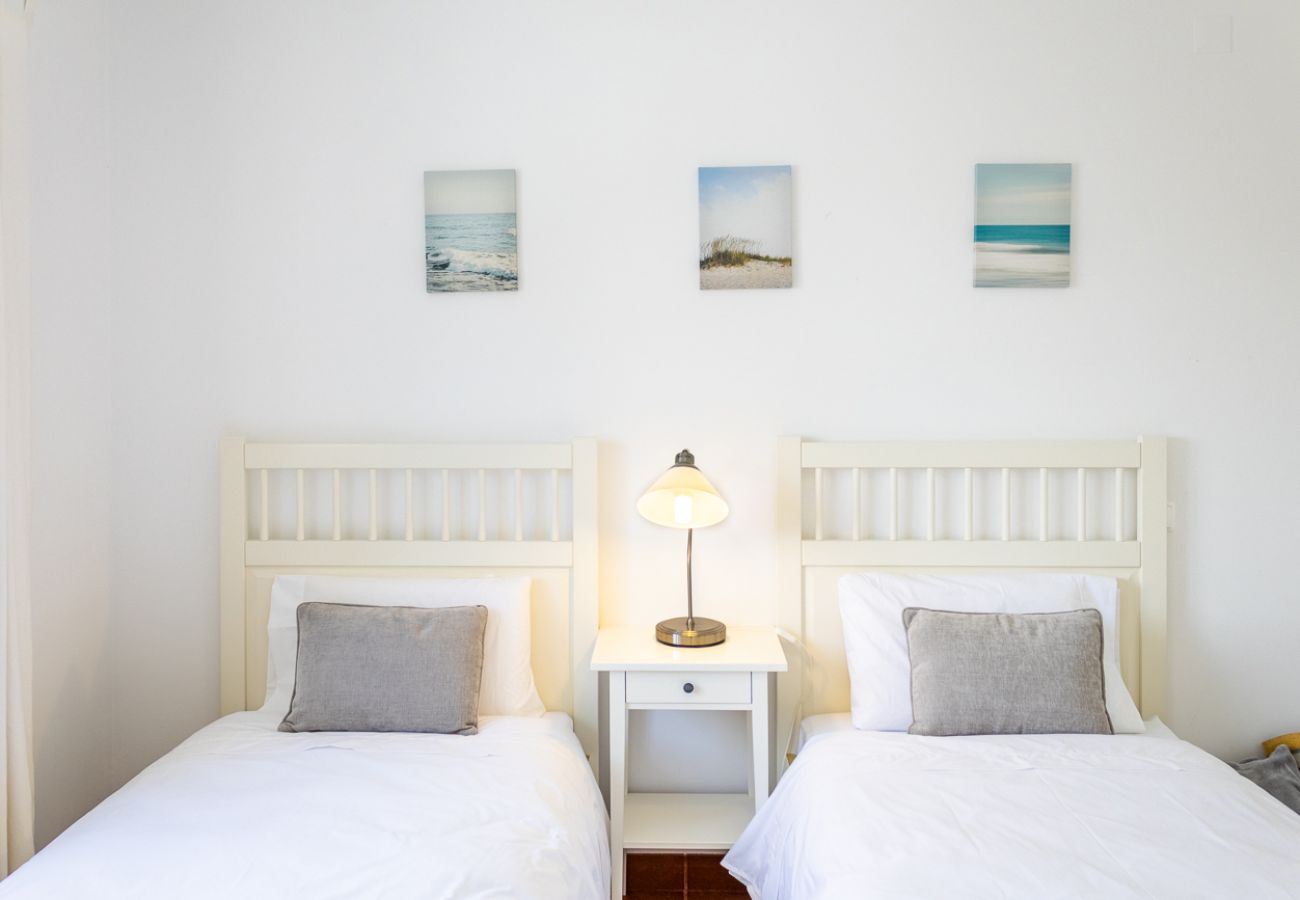 Quarto com duas camas de solteiro, almofadas cinza, mesa de cabeceira branca com abajur e quadros decorativos na parede.