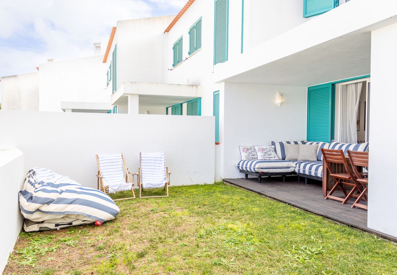 Terraço de uma casa branca com janelas verdes, sofá listrado azul e branco, mesa de jantar de madeira, espreguiçadeiras e pufe em um gramado cercado. 