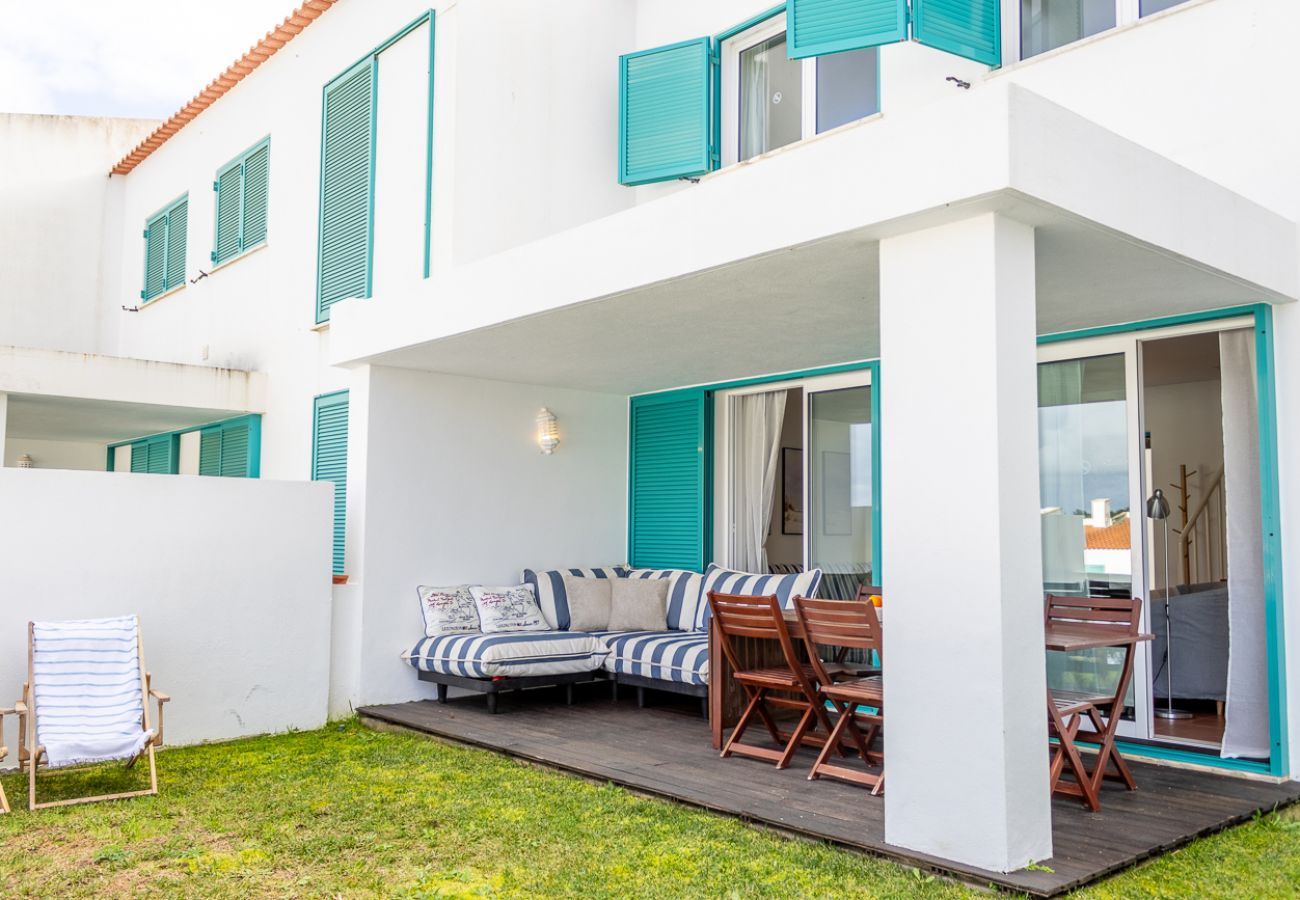 Área externa de uma casa branca com janelas verdes, terraço com sofá listrado azul e branco, mesa de jantar de madeira e gramado com espreguiçadeira.