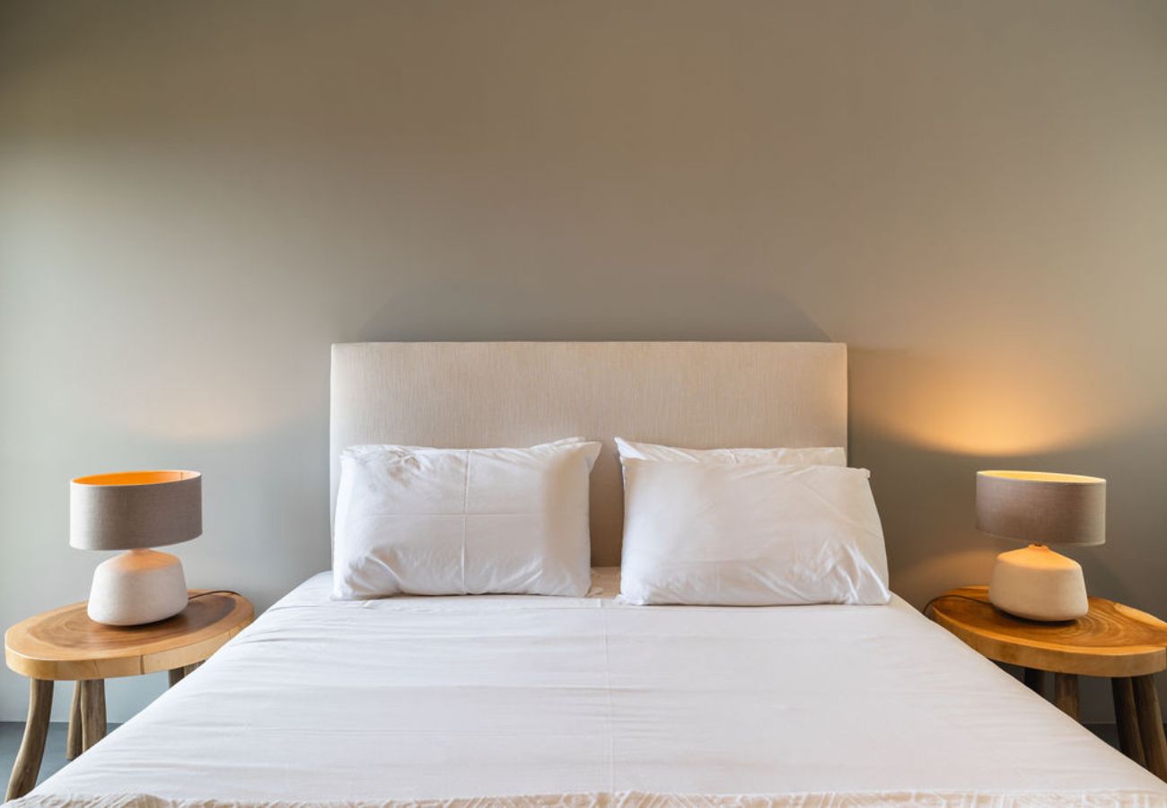 Cama de casal com cabeceira estofada, roupa de cama branca e mesas de cabeceira com luminárias acesas.