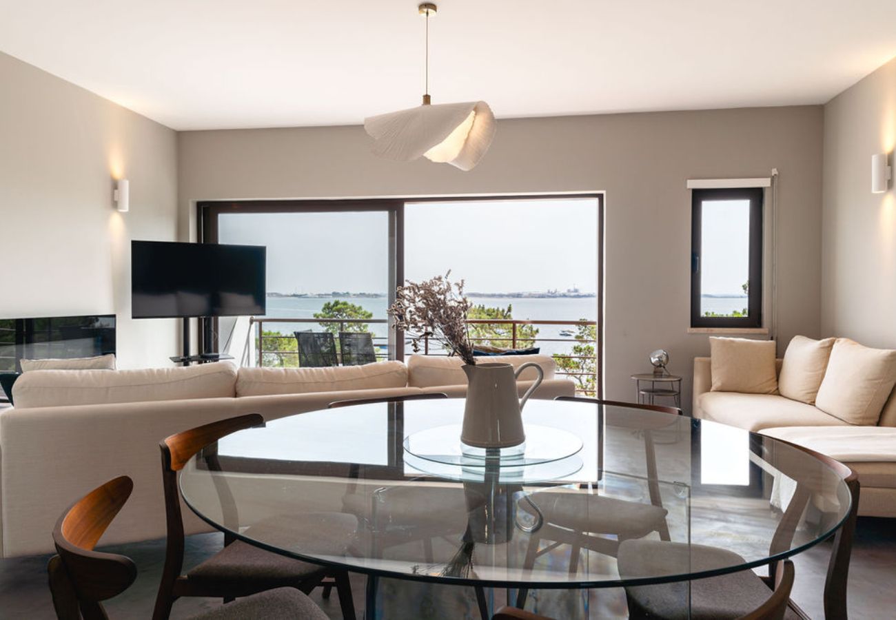 Sala de jantar elegante com mesa de vidro e cadeiras de madeira, integrada a uma sala de estar com sofá bege e vista panorâmica para o mar.
