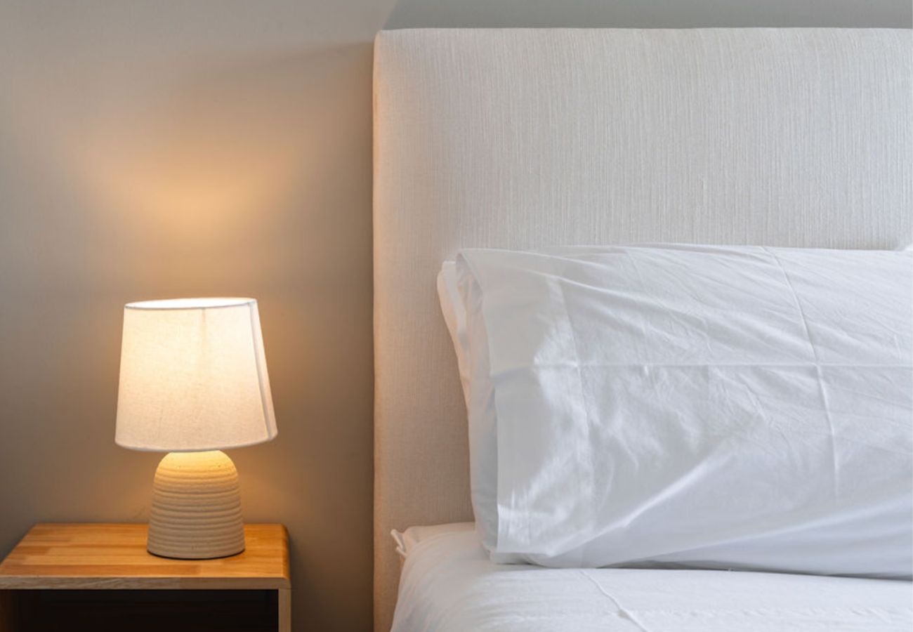 Abajur aceso ao lado de uma cama confortável com cabeceira estofada, criando um ambiente acolhedor.