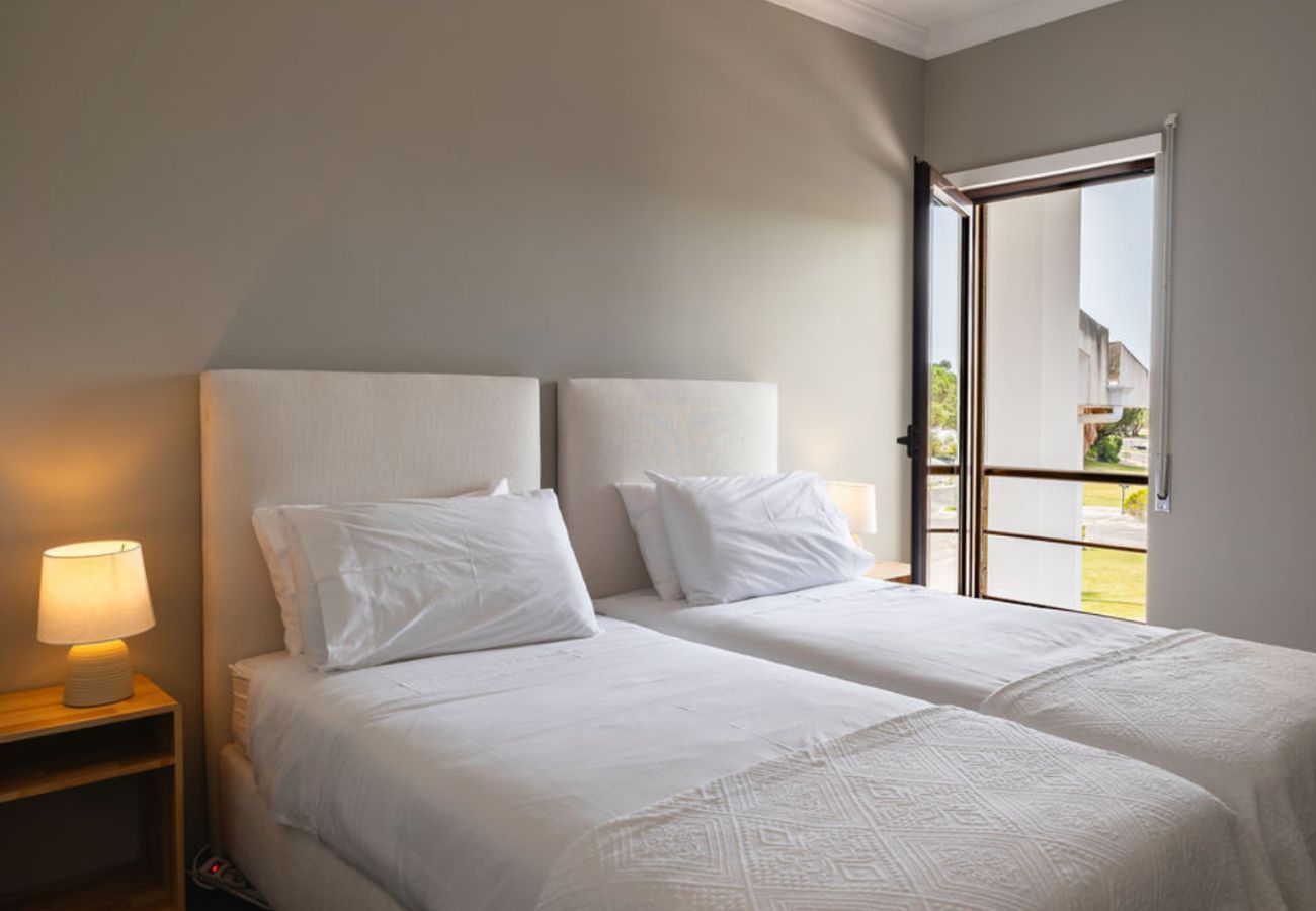 Quarto iluminado com duas camas de solteiro unidas, roupa de cama branca, cabeceira estofada e uma janela ampla com vista externa.