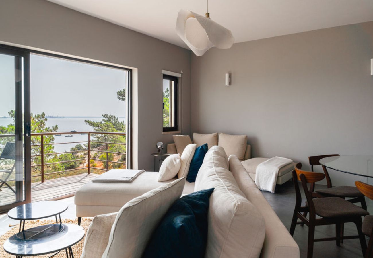 Sala de estar espaçosa com sofás claros, almofadas azuis e varanda com vista para o rio e a vegetação ao fundo.