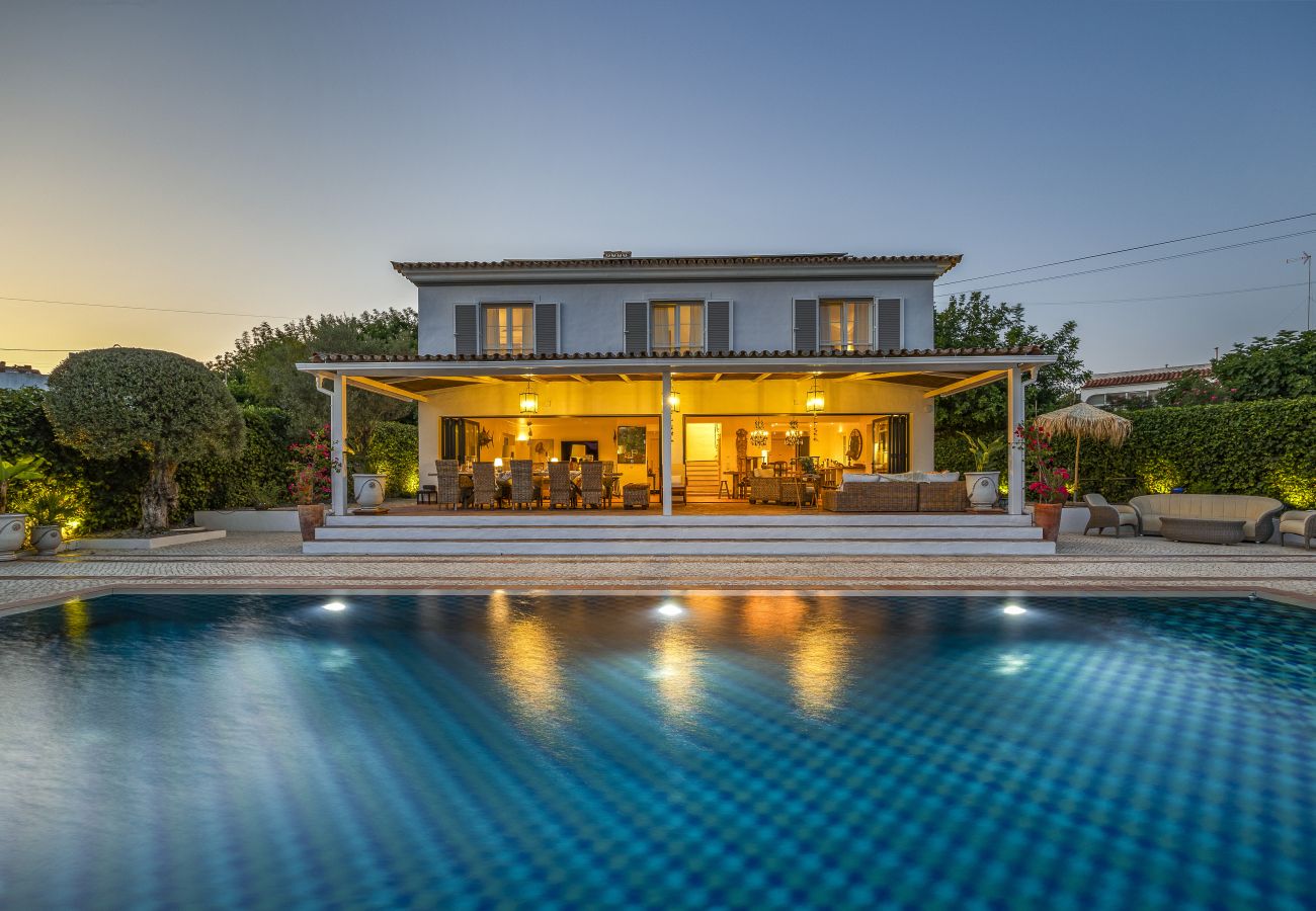 Elegant villa illuminated at sunset, with a pool reflecting the lights and a cozy outdoor area surrounded by greenery.