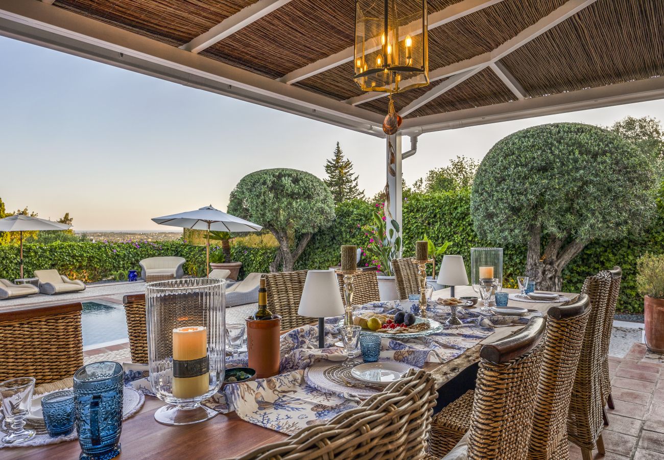 Mesa de jantar externa elegantemente posta, cercada por cadeiras de vime e iluminada por velas, com vista para o jardim e a piscina ao entardecer.