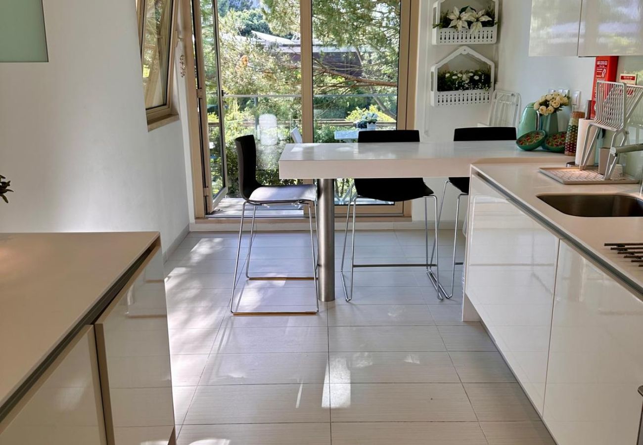 Cozinha elegante com armários brancos, bancadas em vidro e piso claro. Ambiente bem iluminado com acesso a uma área externa através de uma grande port