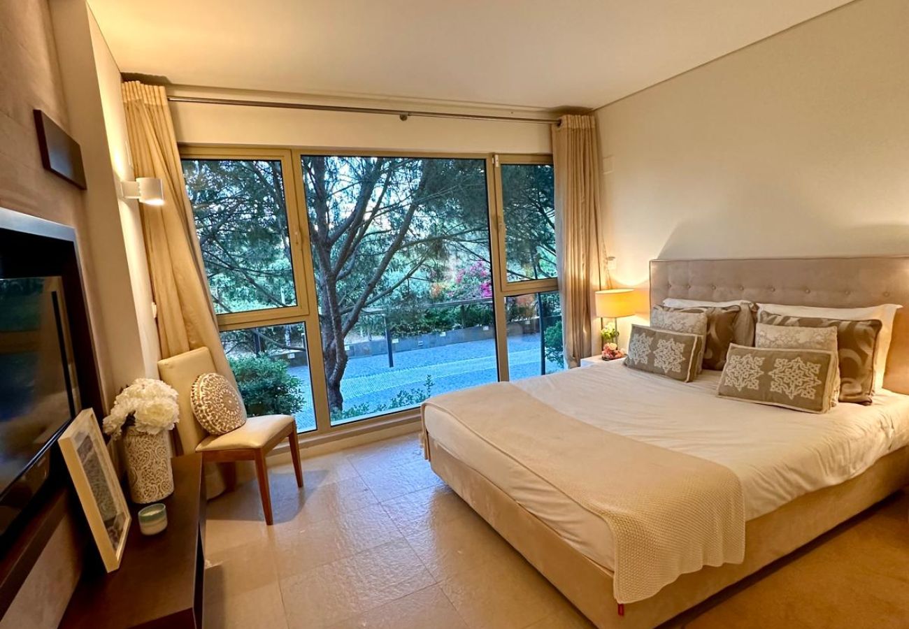 Quarto espaçoso com cama de casal, cabeceira estofada e iluminação suave. Grandes janelas de vidro oferecem vista para o jardim, permitindo a entrada 