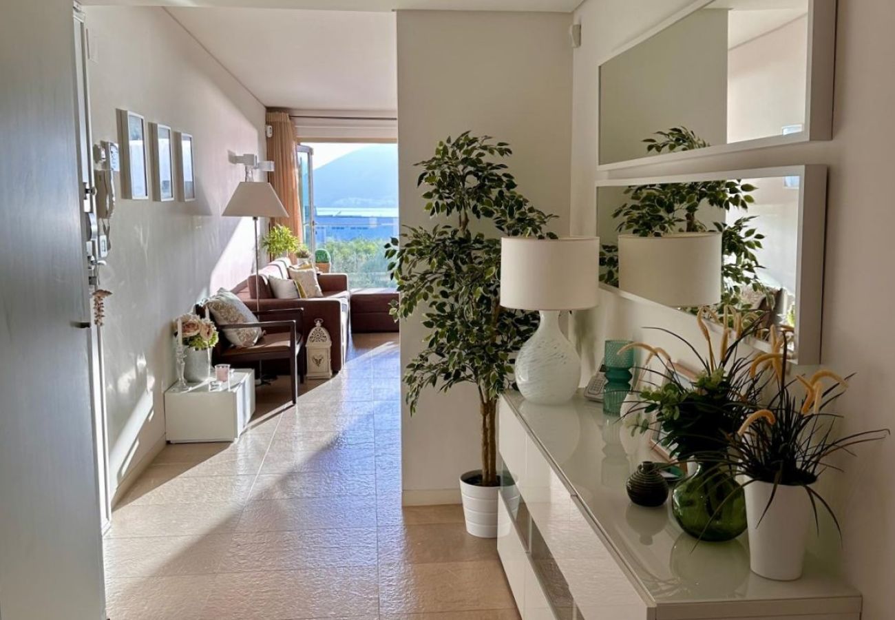 Corredor iluminado com piso de pedra clara, móvel branco decorado com vasos de plantas e espelhos na parede. Ao fundo, sala de estar com sofá e vista 