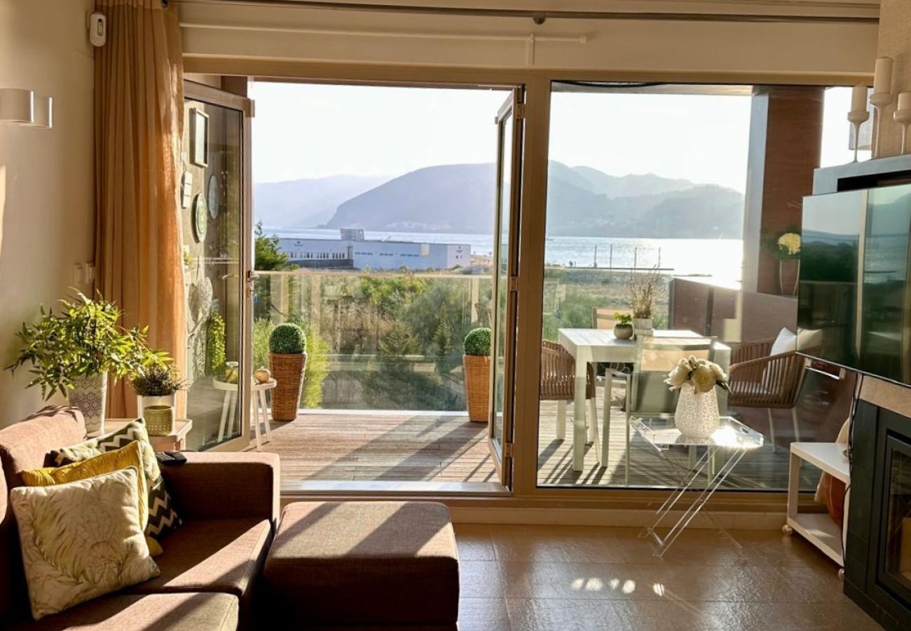 Sala de estar moderna e iluminada, com sofá confortável e grandes portas de vidro que levam a um terraço com vista para o mar e montanhas.