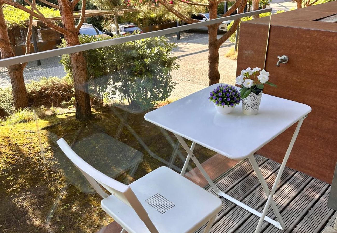 Terraço compacto com piso de madeira, mesa e cadeiras brancas dobráveis. Pequenos vasos de flores decoram o espaço, que tem vista para uma área arbori