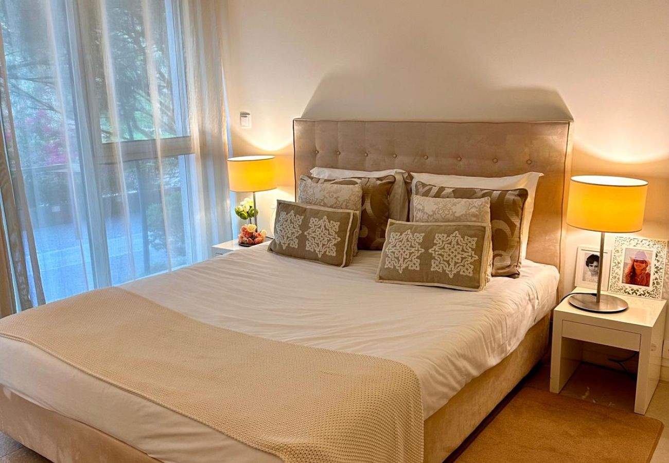 Quarto espaçoso com cama de casal, cabeceira estofada e iluminação suave. Grandes janelas de vidro oferecem vista para o jardim, permitindo a entrada 