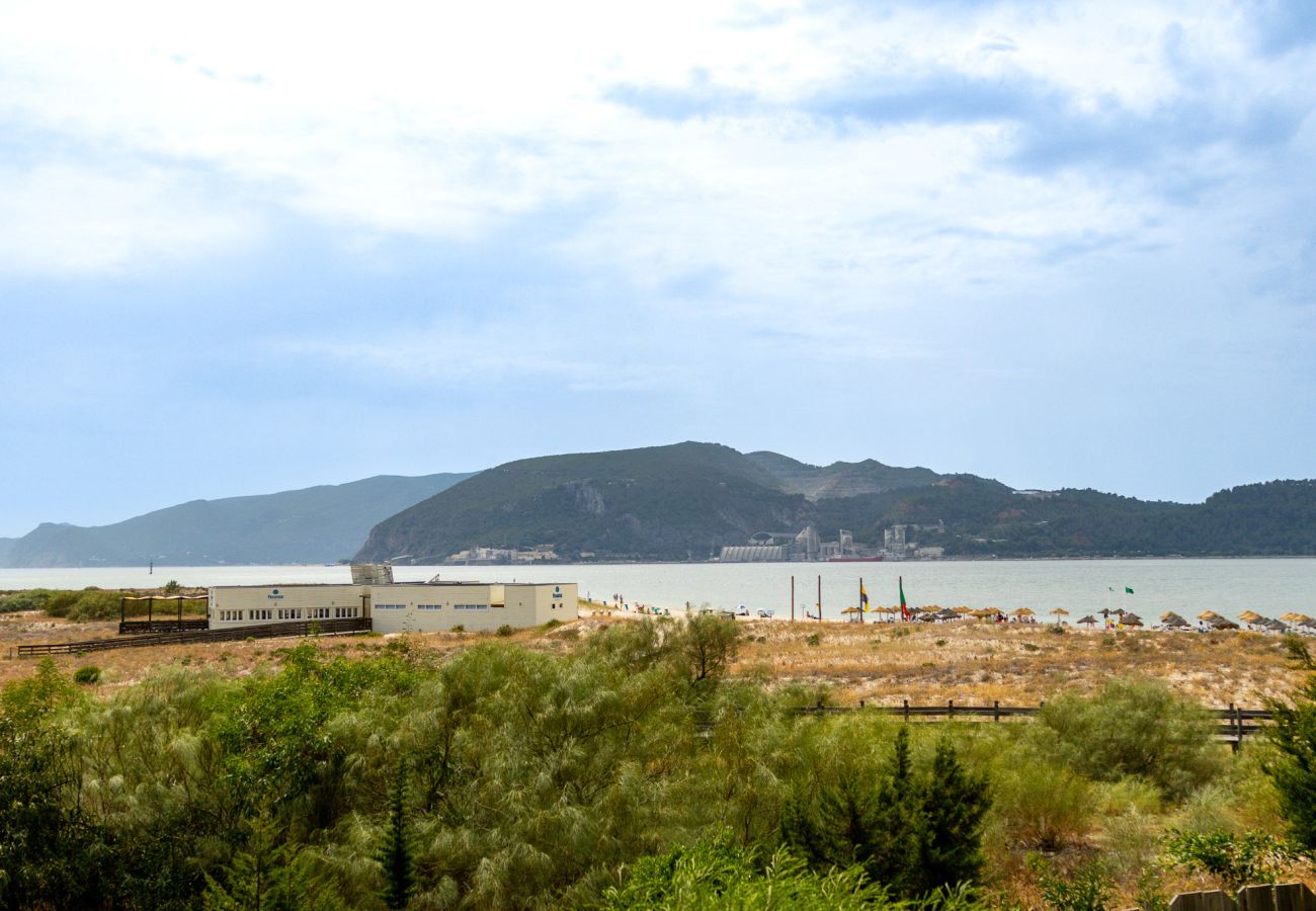 Vista aberta para uma paisagem natural com vegetação verde, um edifício à beira-mar e uma praia com guarda-sóis ao fundo.