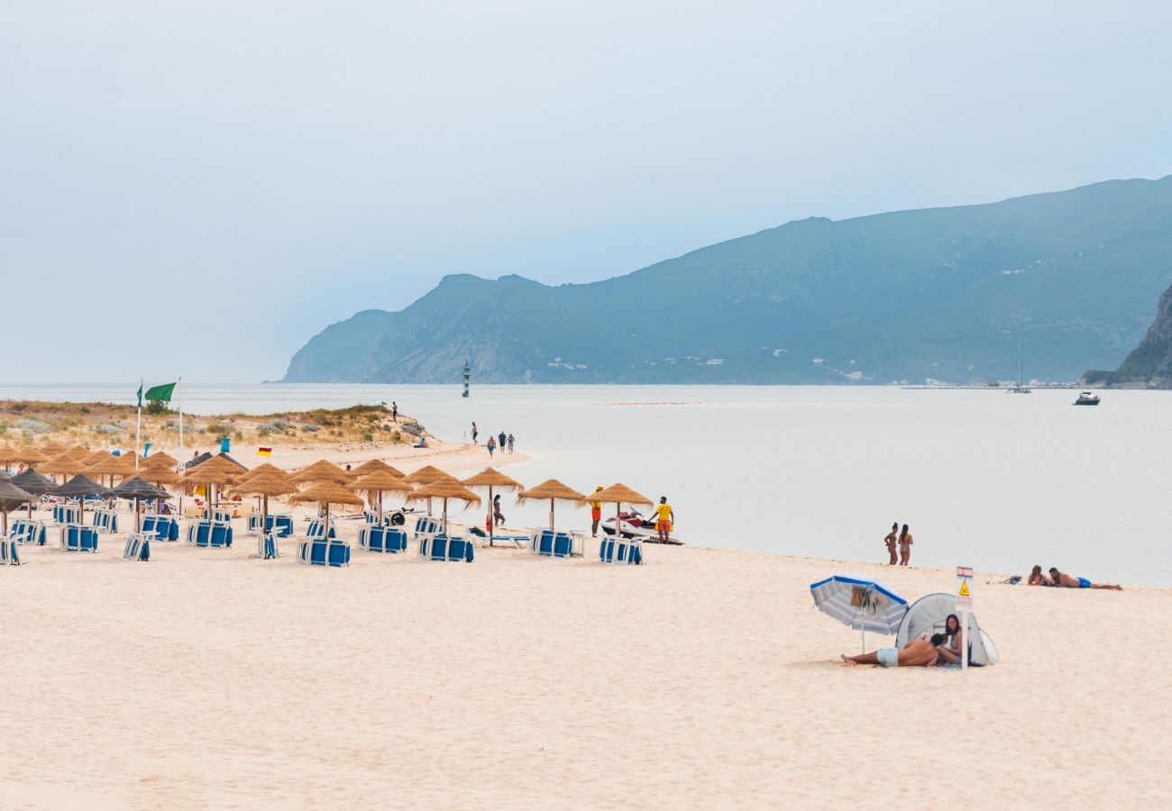 Areia dourada, mar sereno e guarda-sóis de palha organizados na praia de Tróia. Cenário relaxante com montanhas ao fundo e algumas pessoas desfrutando