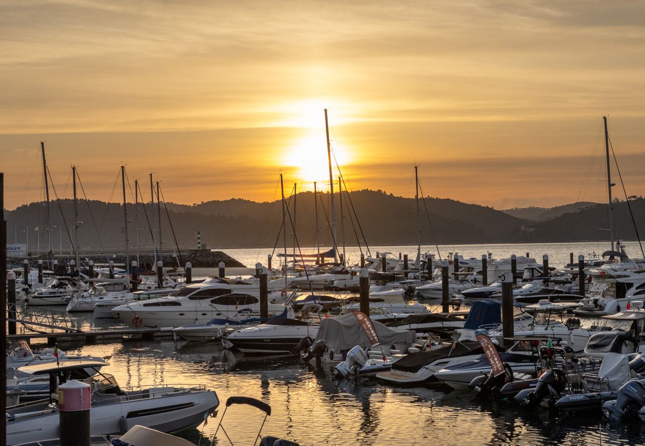 Uma marina repleta de iates e barcos ao pôr do sol, com o reflexo dourado da luz sobre a água e montanhas ao fundo.