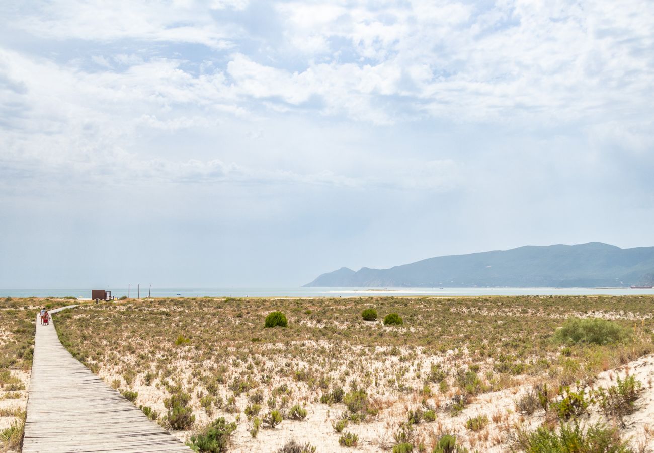 Um caminho de madeira atravessa uma área de dunas com vegetação rasteira, levando até a praia e o mar ao fundo, com montanhas e céu parcialmente nubla