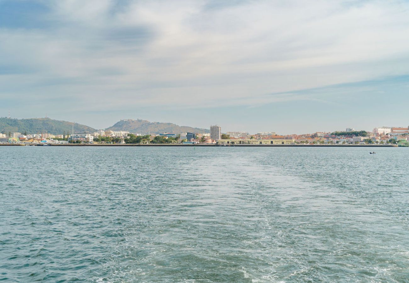 Vista da água azul com ondulações e rastro de barco, com uma cidade e montanhas ao fundo sob um céu parcialmente nublado.