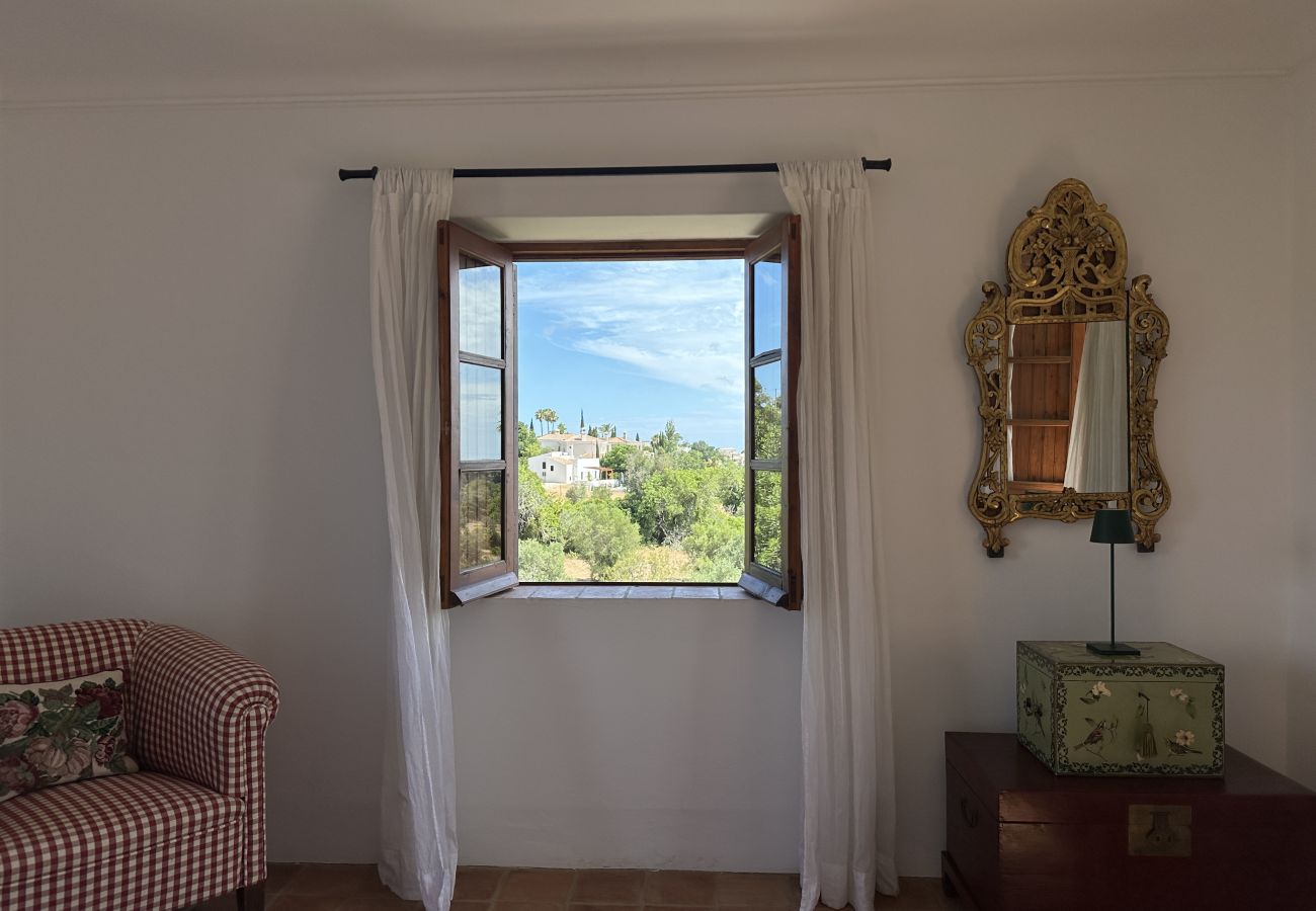 Quarto decorado com poltrona xadrez, espelho dourado e uma janela aberta revelando uma paisagem verde exuberante.