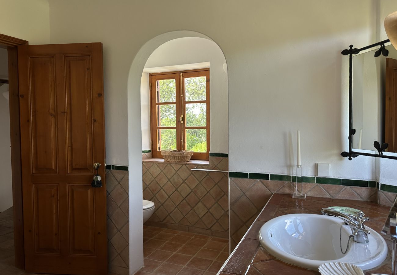 Banheiro espaçoso com pia de cerâmica branca, espelho com moldura de ferro, azulejos rústicos e uma charmosa janela de madeira com vista para a nature