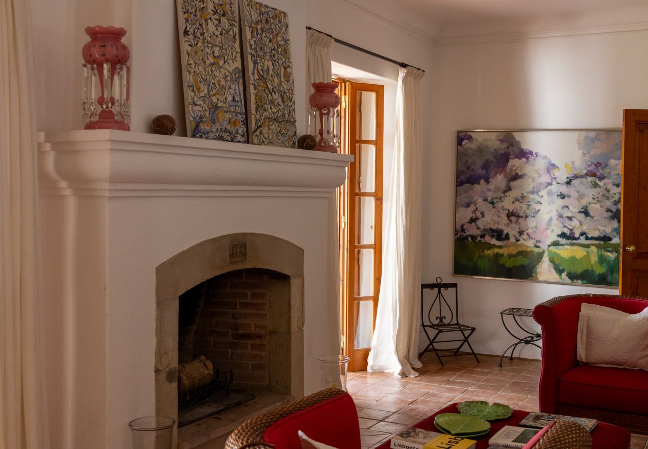 Sala de estar com lareira branca e detalhes em azulejo, sofá vermelho com almofadas, mesa de centro com livros e objetos decorativos, e um quadro colo