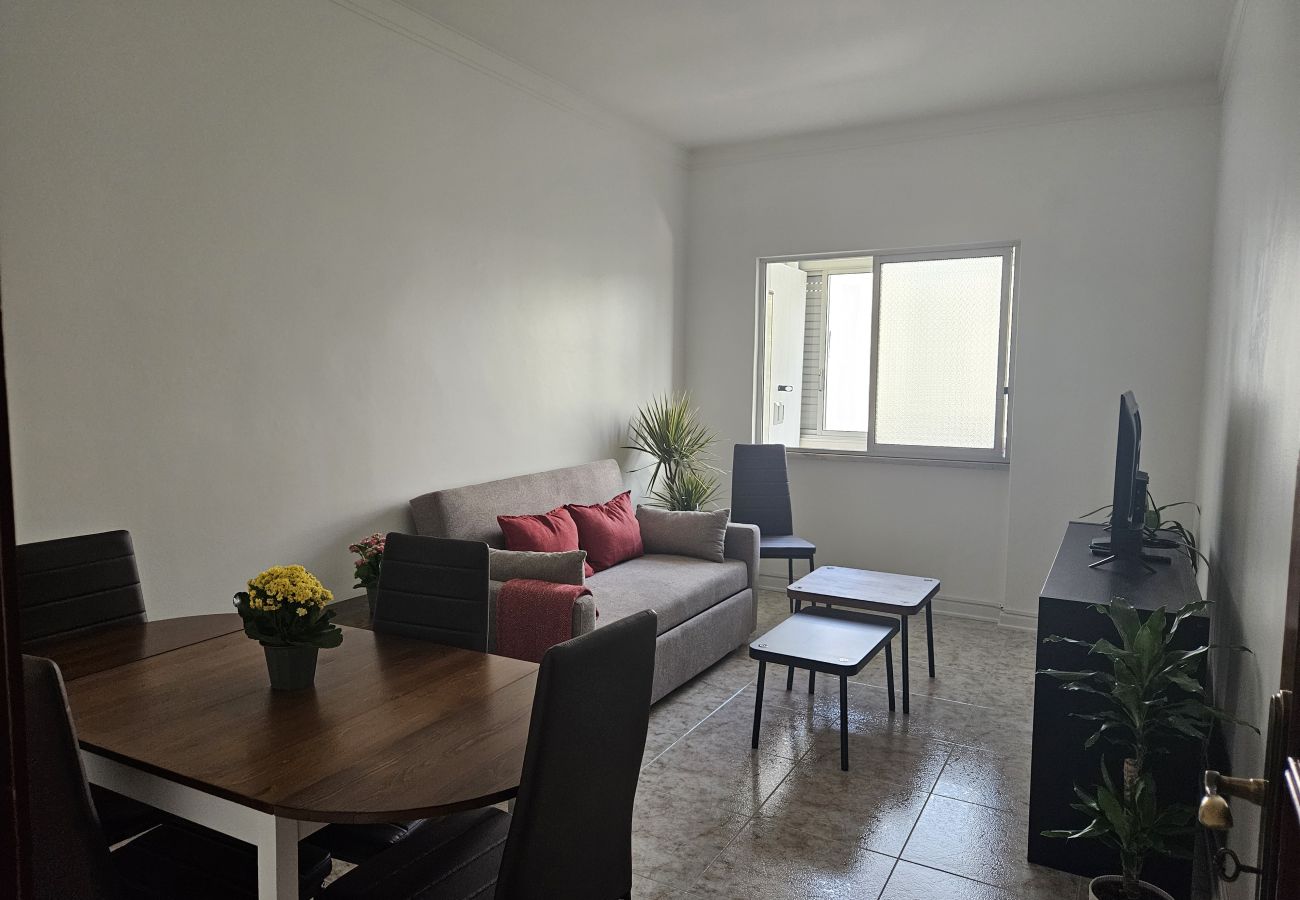 Sala de estar iluminada com mesa de jantar de madeira, cadeiras escuras, sofá com almofadas vermelhas, televisão e plantas decorativas.