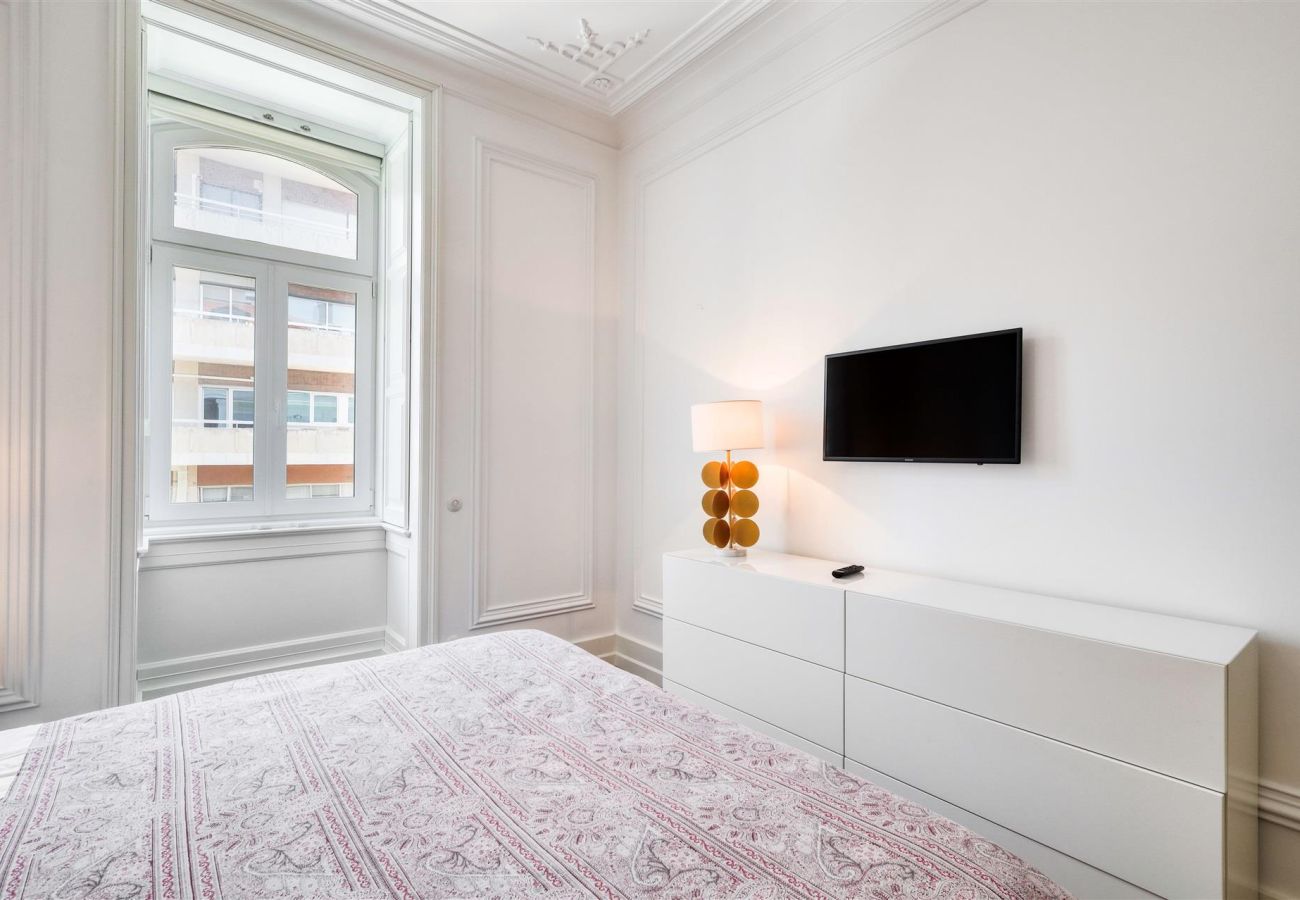 Quarto espaçoso com cama de casal, cômoda branca, TV de tela plana e grande janela para iluminação natural.