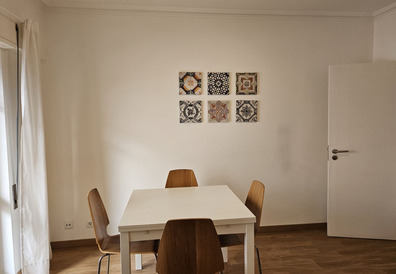 Dining room with a white table and wooden chairs, decorative tile art on the wall, and light curtains allowing natural light.