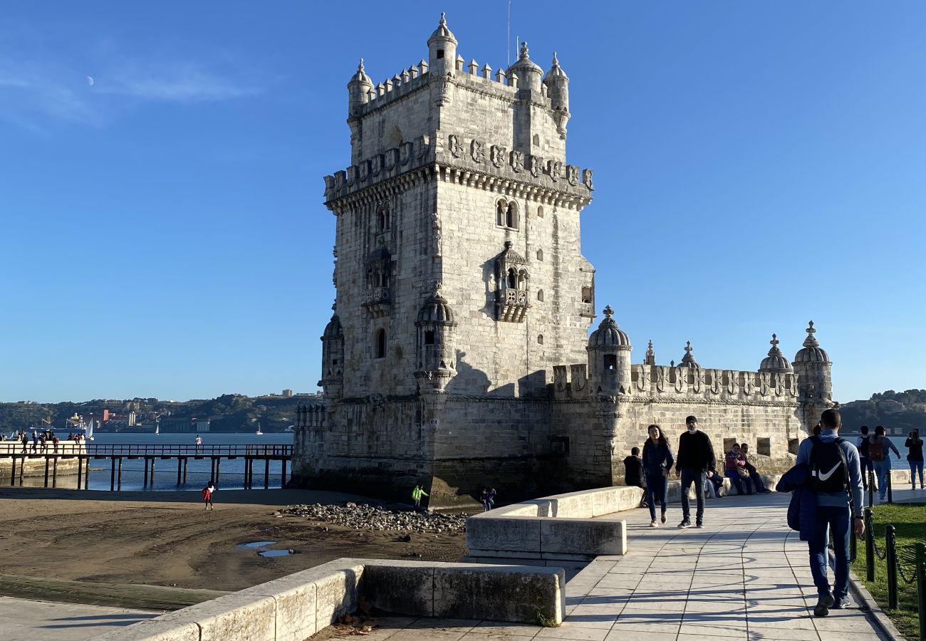 Belém Tower by the Tagus River, with tourists walking along the promenade on a sunny day.