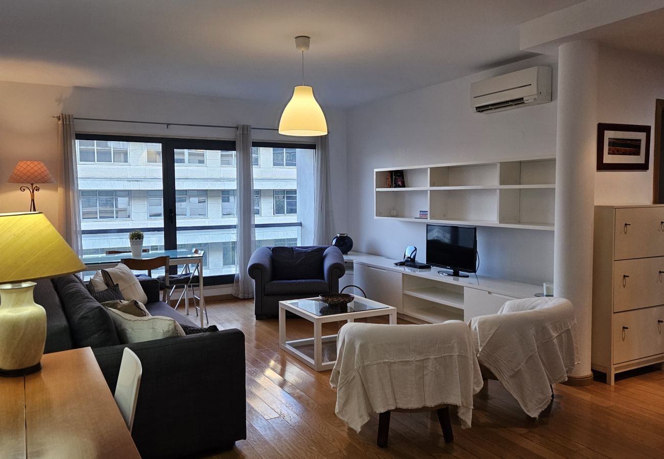Modern living room with a sofa, armchairs, white shelving unit, TV, and large windows with an urban view.