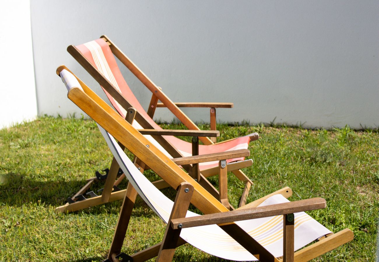 Two wooden lounge chairs with striped fabric positioned on the grass, with a white wall in the background and houses in the distance.