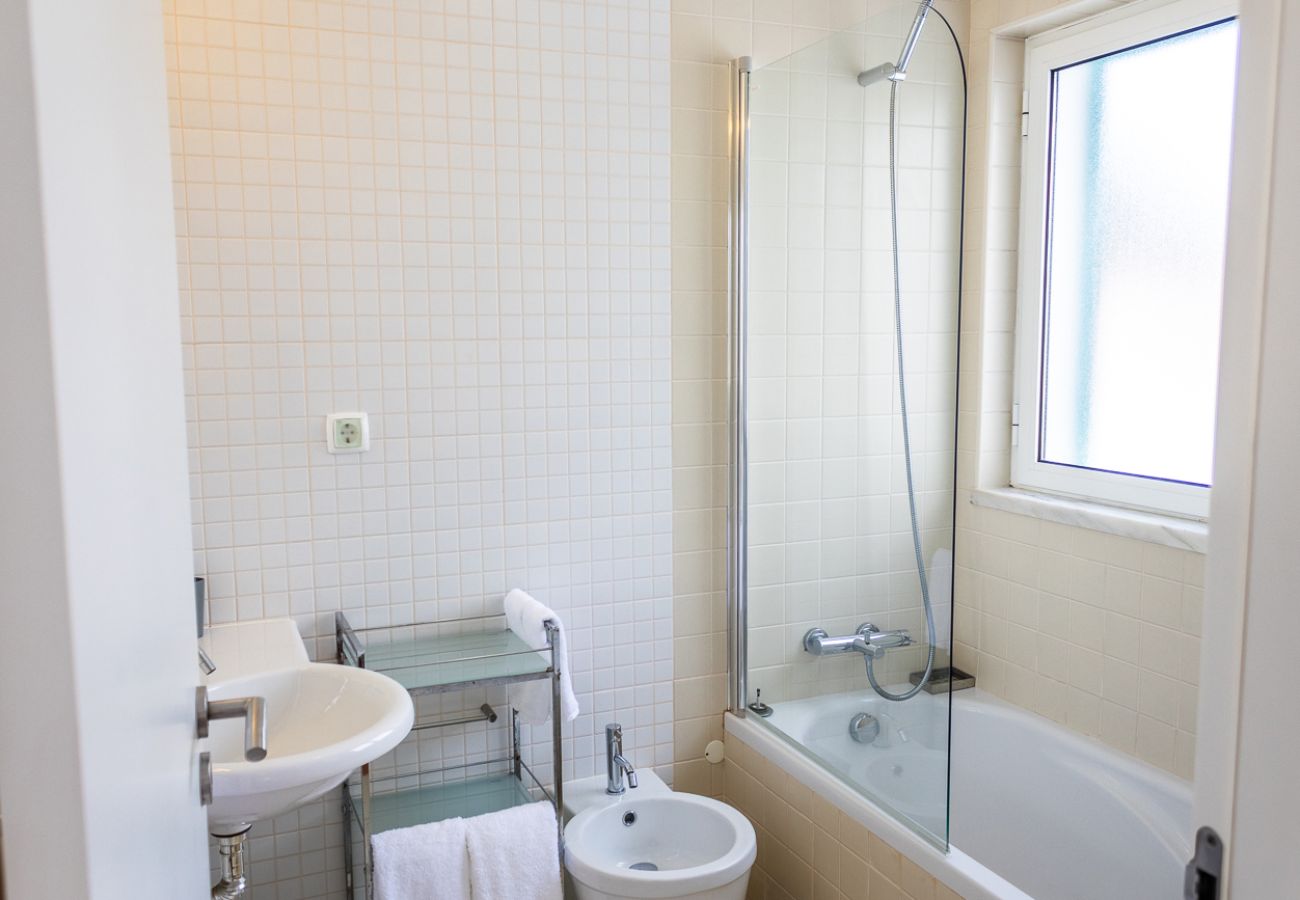Bathroom with a glass bathtub, white sink, bidet, and white towels, featuring a large window allowing natural light.