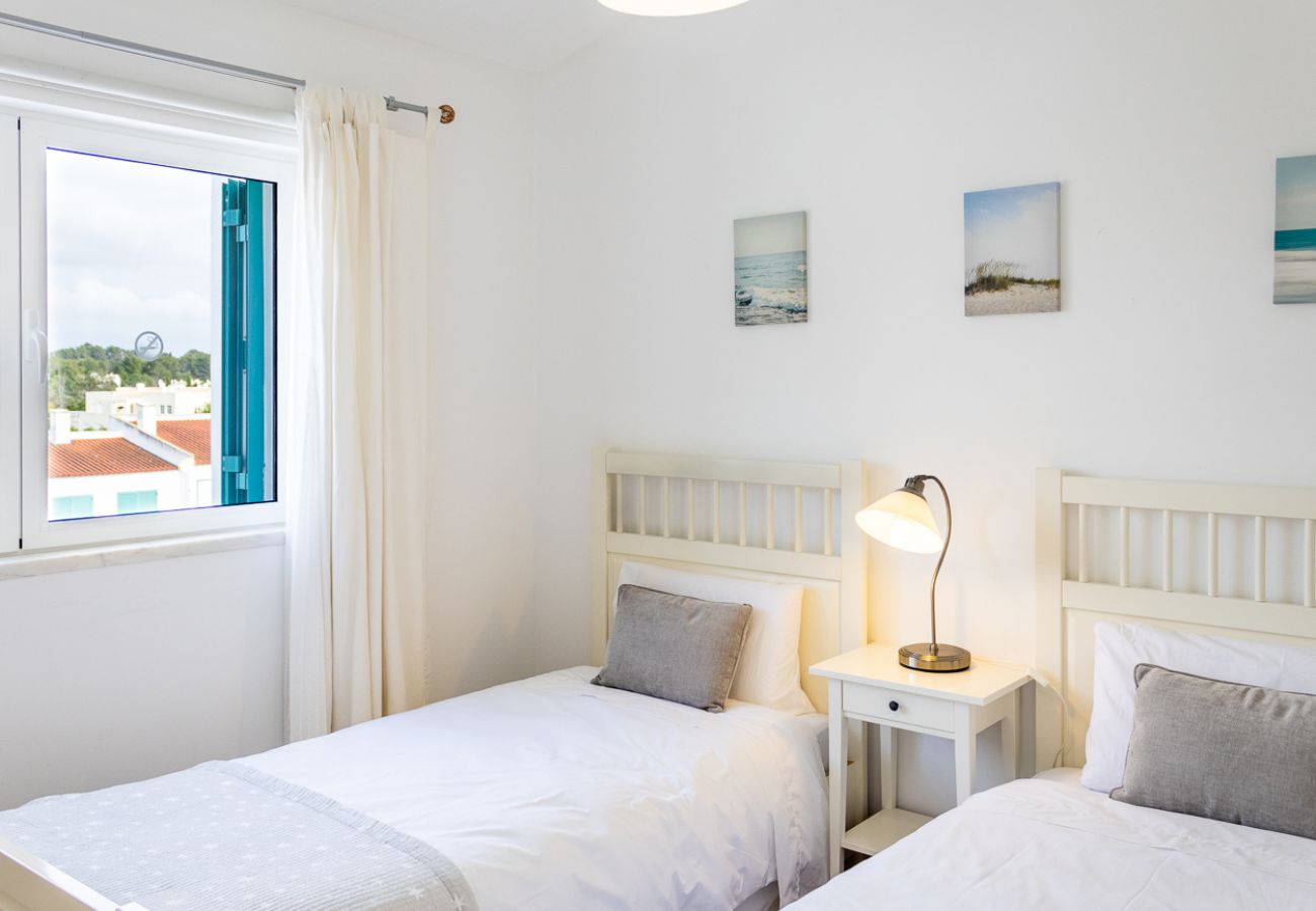 Bedroom with two single beds, gray pillows, bedside table with lamp, and large windows with white curtains.