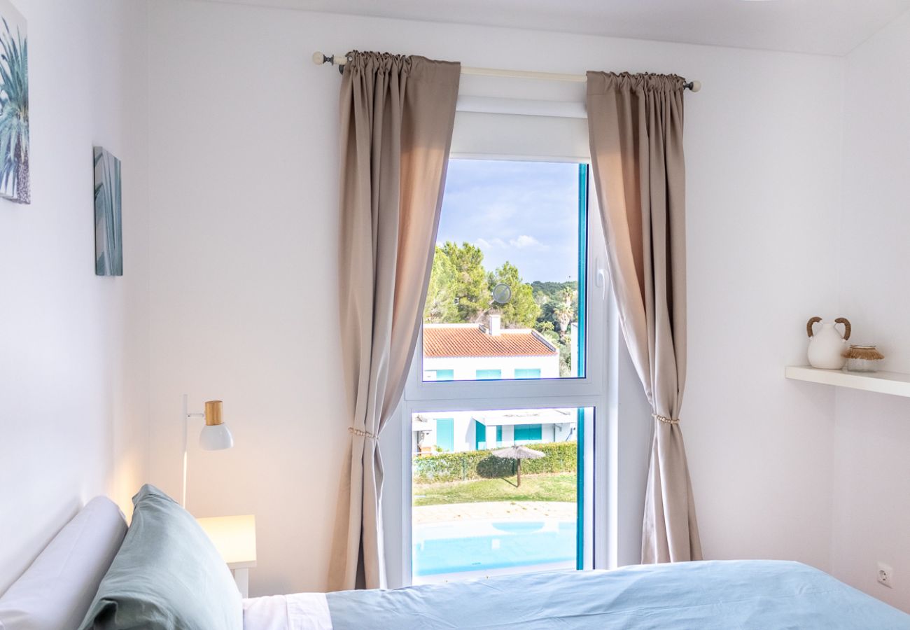 Bedroom with a double bed, beige curtains, and a large window overlooking the pool and green area.