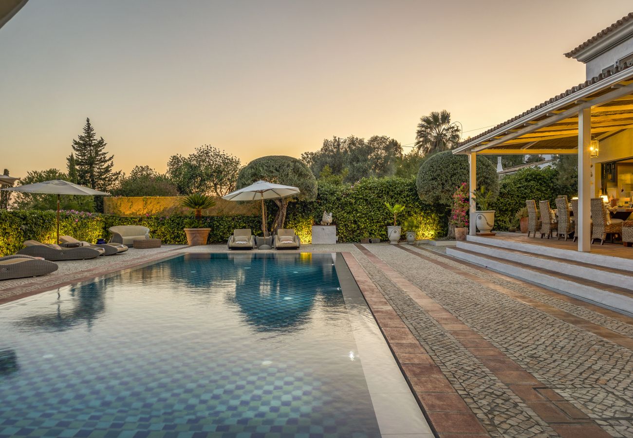  Pool reflecting the golden sunset sky, surrounded by sun loungers and umbrellas, with the softly lit outdoor dining area in the background.