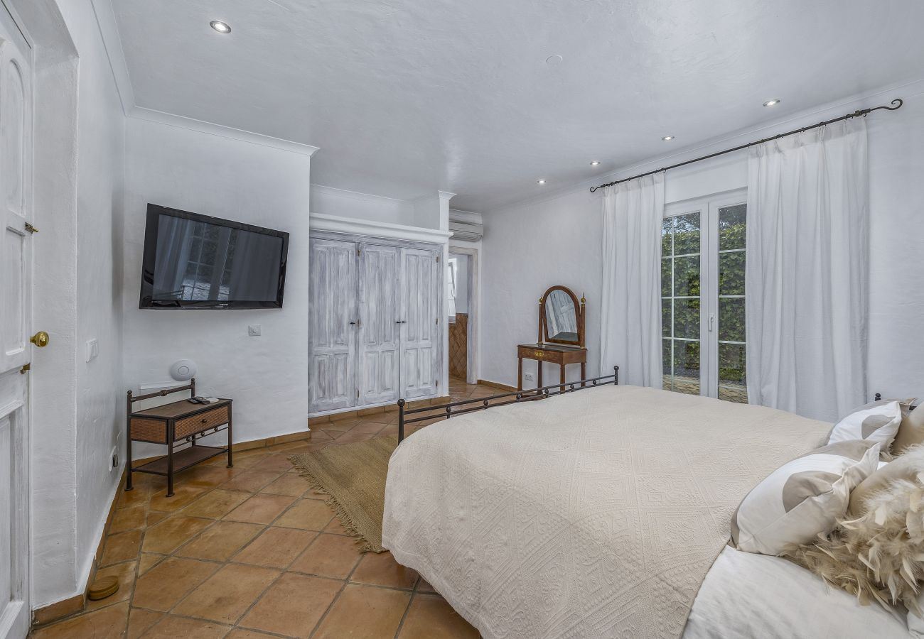 Spacious bedroom with a double bed, wall-mounted TV, elegant wardrobes, and a large window with white curtains overlooking the garden.