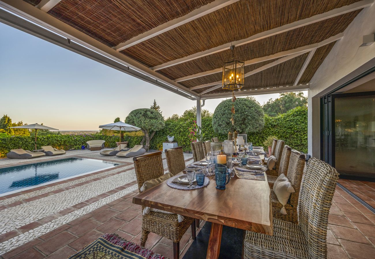 Elegantly set outdoor dining table under a rustic pergola, overlooking the pool with a serene sunset atmosphere.