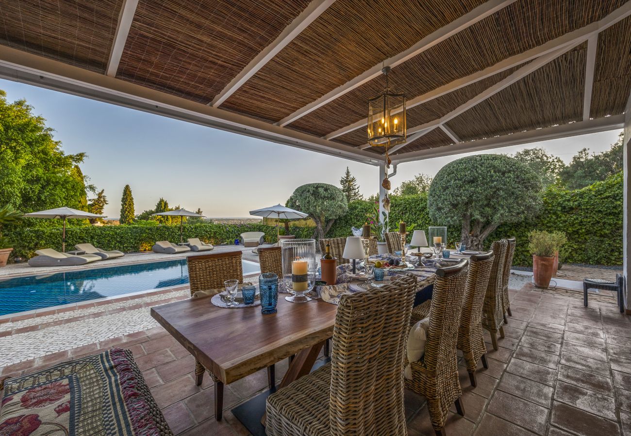 Covered outdoor dining area with an elegantly set table under a straw roof, pool view, and warm lighting.