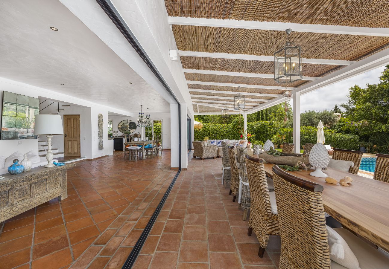 Spacious covered outdoor area with a rustic dining table, wicker chairs, and views of the pool and garden.