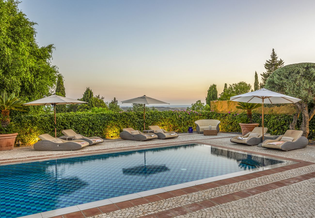 Piscina de azulejos reflejando el cielo al atardecer, rodeada de tumbonas cómodas y sombrillas entre vegetación exuberante.