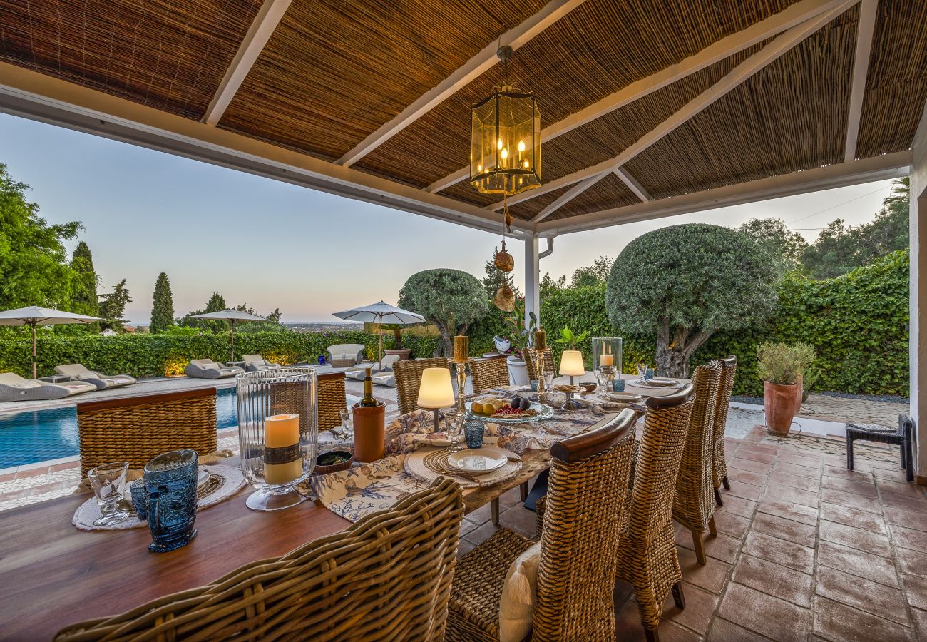 Covered outdoor dining area with an elegantly set table under a straw roof, pool view, and warm lighting.