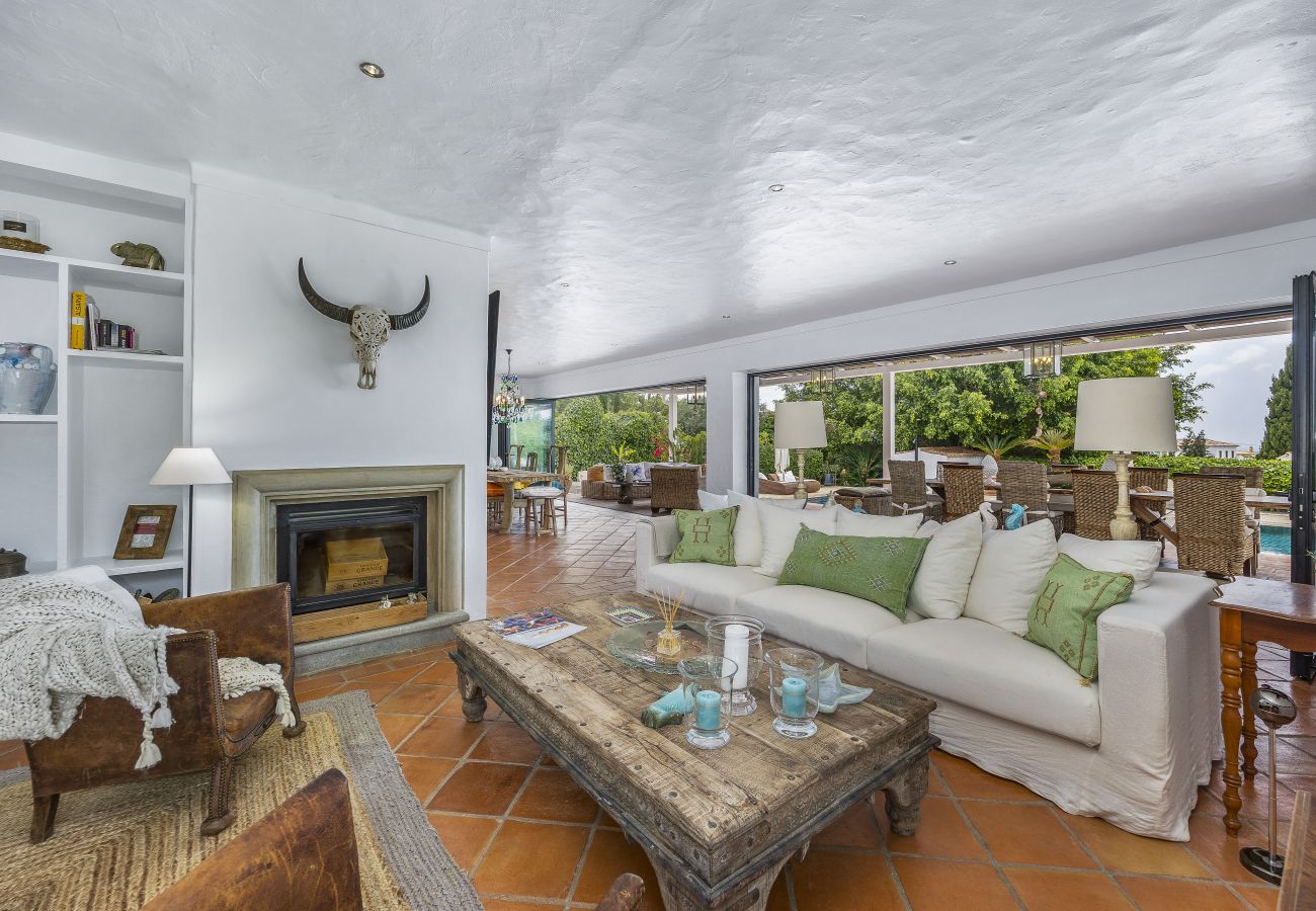 Cozy living room with white sofas, a fireplace, a rustic wooden table, and large windows opening to a lush terrace.