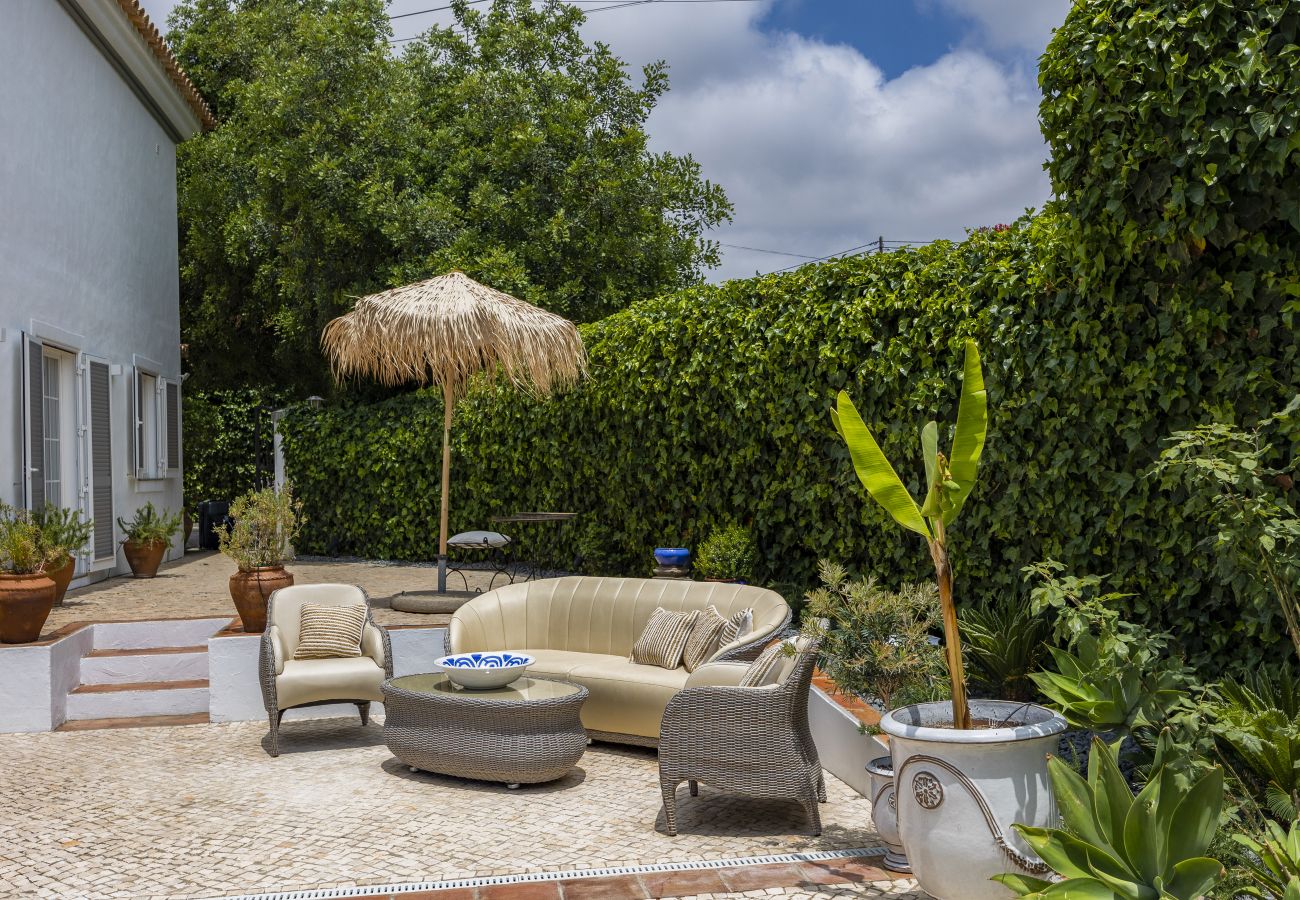 Cozy outdoor space with comfortable sofas and armchairs, surrounded by a green wall covered in plants and a straw parasol.