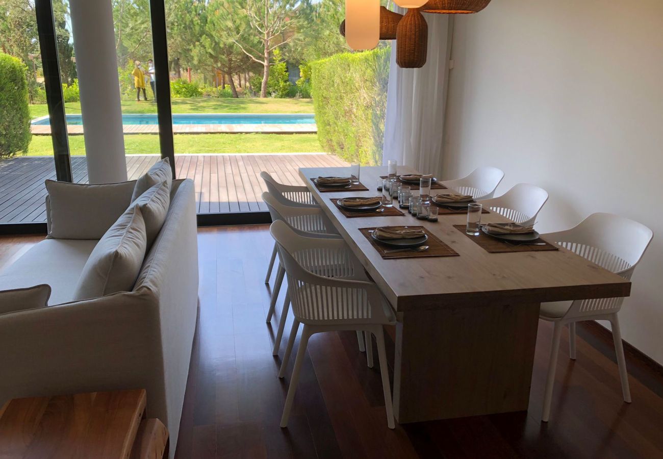 Elegant dining table set for meals, featuring white chairs, pendant lighting, and a panoramic view of the pool and garden.